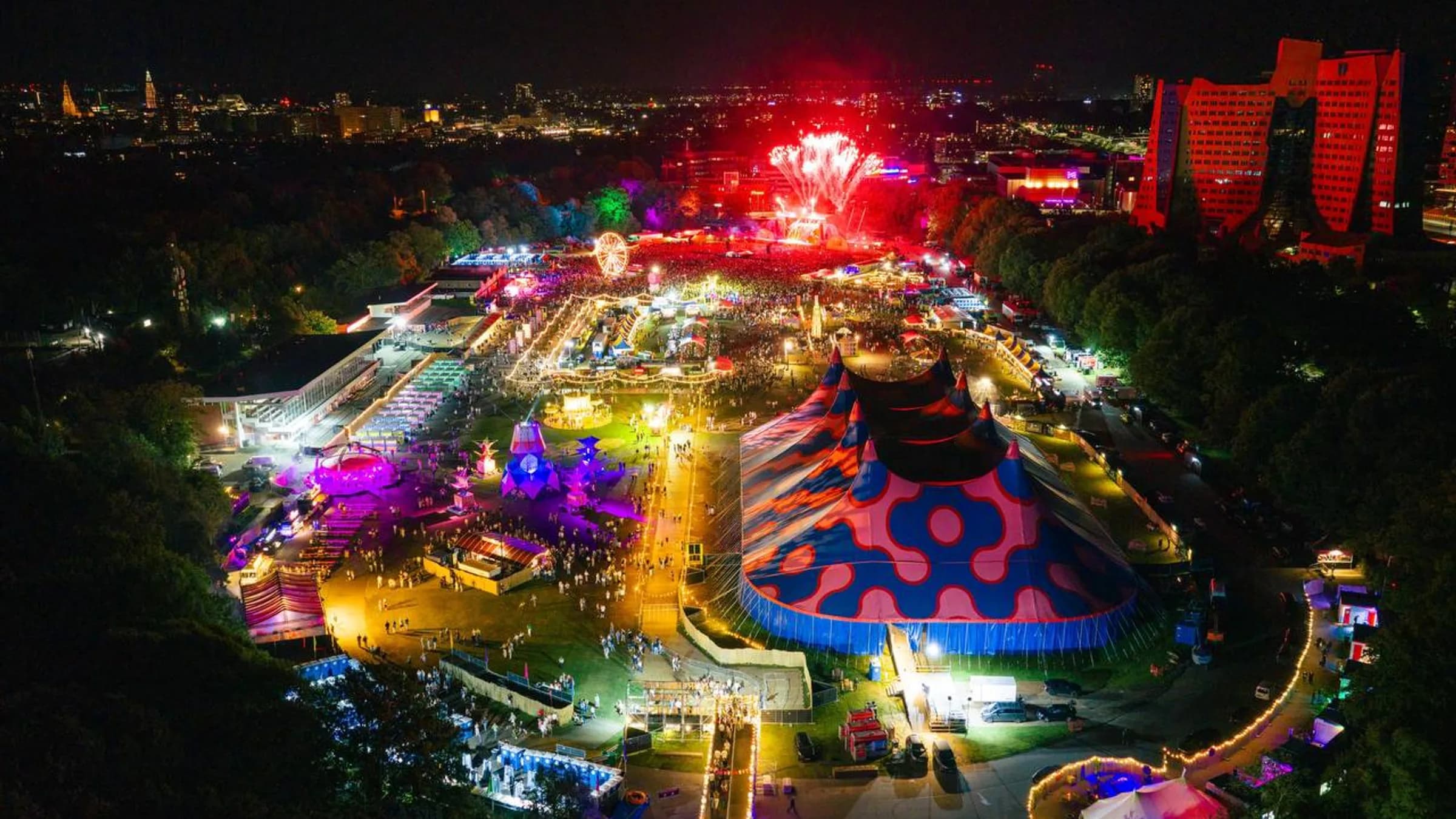 Luchtfoto van Hullabaloo bij avond, met de verlichte tent, het festivalterrein in het park en de skyline van Groningen op de achtergrond